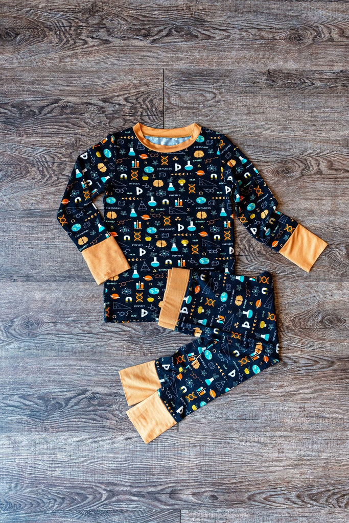 Keep an Ion Bedtime Big Kids Jams black pajama set with a colorful science-themed pattern includes DNA helixes, atoms, microscopes, flasks, beakers, test tubes, mathematical equations, books, the word 'Physics', and the word 'Chemistry'. There are also icons of planet earth,ring planet evolution of moon, light bulbs, and scientific symbols in colors such as orange, blue, green, and white. The pajama top has long sleeves with orange cuffs and trim matching orange neckline on a wooden floor background.