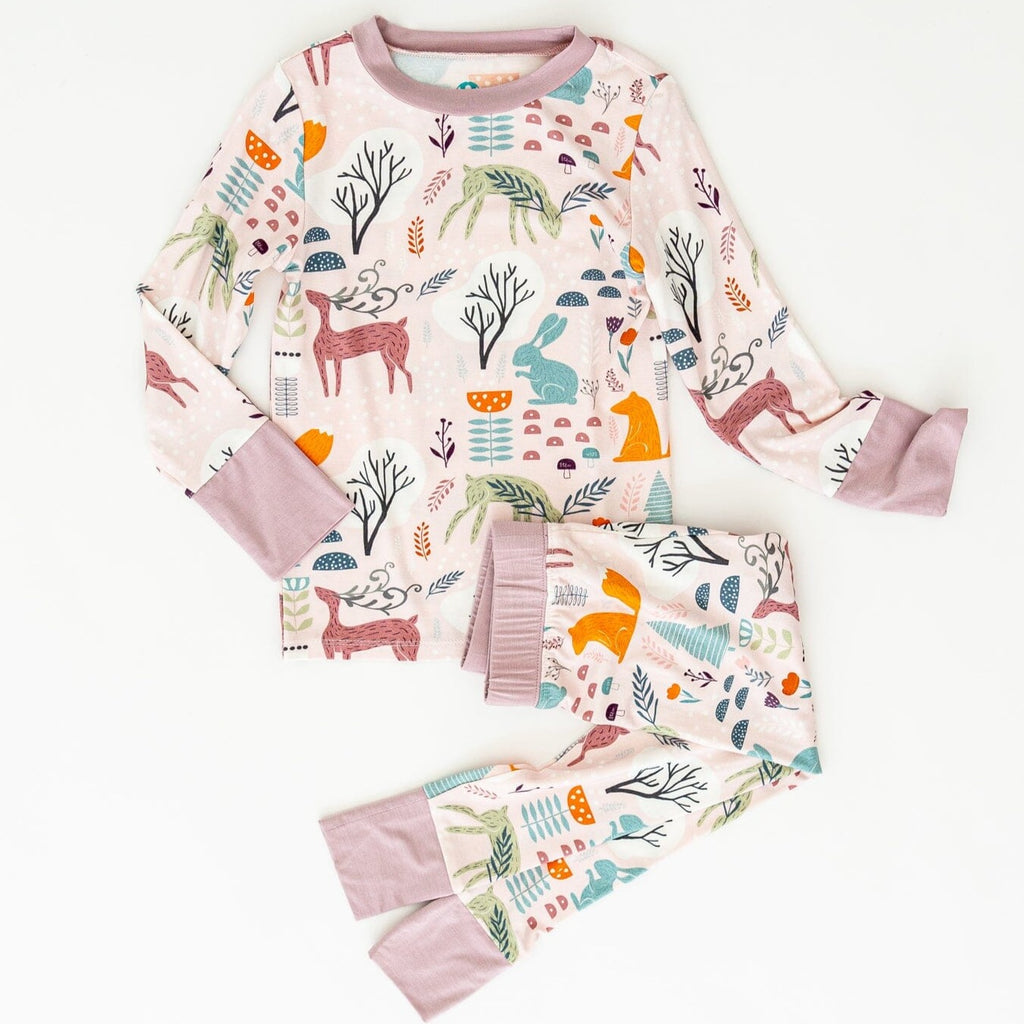 Meadowlark Big Kid Jams a light pink pajama set featuring a whimsical woodland pattern. The design includes various animals such as deer, rabbits, and foxes, along with trees, mushrooms, and foliage in shades of green, orange, blue, and brown. The pajama has light pink cuffs and a matching neckline, laid out against a plain white background.