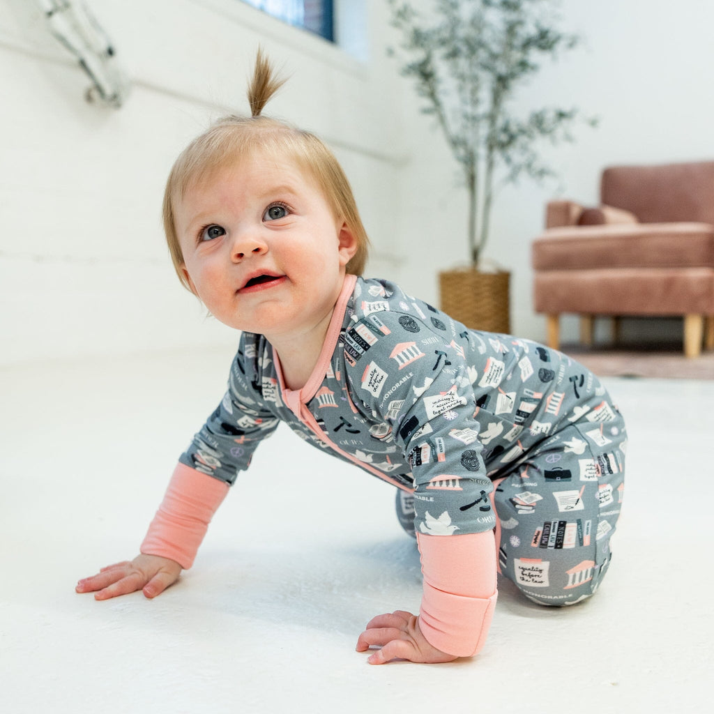 Order in the Cot Jams a young girl with light brown hair styled in a small ponytail on top is crawling on a white floor. The she is wearing a grey footie pajama with law-themed illustrations, including scales of justice, gavels, law books, and courthouse buildings. The pajama features light pink cuffs and a light pink two-way zipper running down the front. The child looks up with a curious expression. In the background is a light pink upholstered light pink chair, a woven basket with a plant.