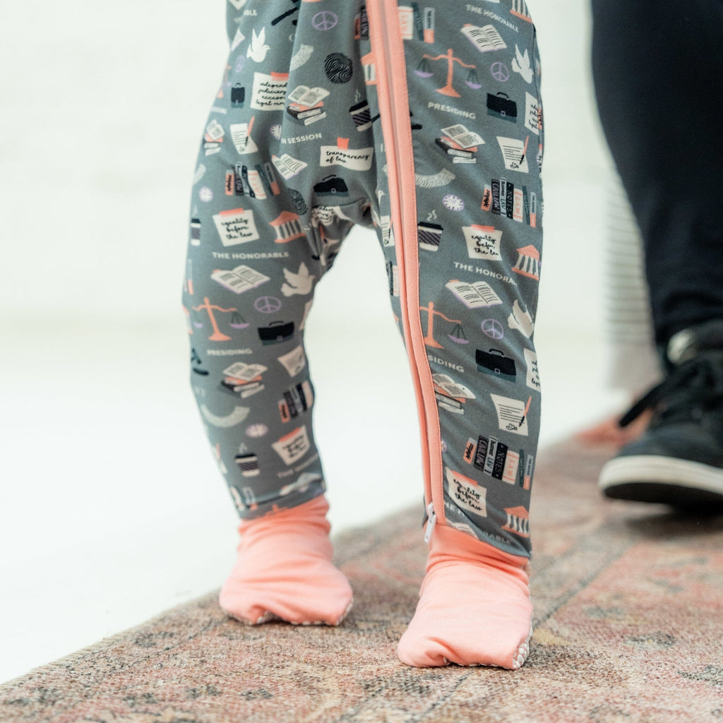 Order in the Cot Jams close-up of a toddler's legs and feet, showing them standing on a carpeted surface. The child is wearing a grey footie pajama with law-themed illustrations, including scales of justice, gavels, law books, courthouse buildings, and phrases like 'The Honorable' and 'Presiding.' The pajama has a pink two-way zipper running down the front and light pink fold-over footies. In the background is an adult's legs and feet, wearing black pants and black shoes.