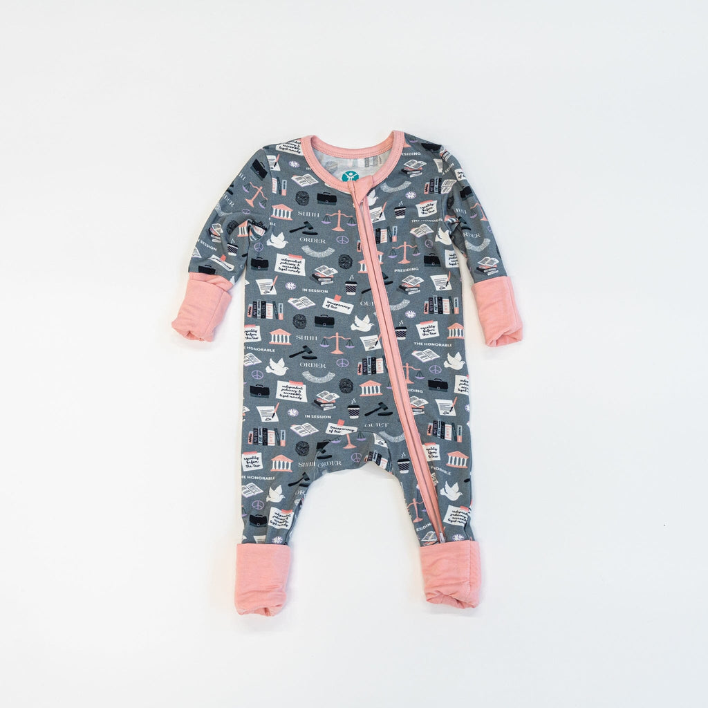 Order in the Cot Jams a grey baby footie pajama with a colorful pattern featuring various legal-themed items. The pattern includes books, gavels, scales of justice, courthouse icons, law symbols, and documents with text that reads 'LAW', 'ORDER', 'CRIME', 'SHERIFF', and 'PRISON'. The footie pajama has a two-way zipper running down the front, light pink trim, and light pink cuffs.