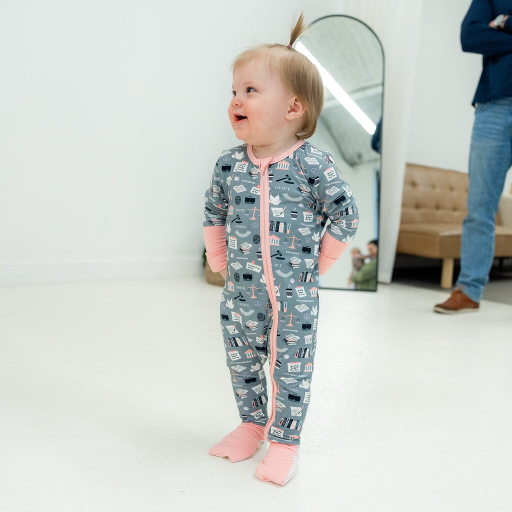 Order in the Cot Jam young girl with light brown hair in a small ponytail on top smiles as she stands and looks to the right. She wears a grey footie pajama with law-themed illustrations like scales of justice, gavels, law books, and courthouse buildings. The pajama features light pink cuffs, trim, and a pink two-way zipper. Her hands are playfully tucked inside the sleeves. The background includes a large arched mirror, a light pink tufted sofa, and an adult partially out of frame in jeans and brown shoes.