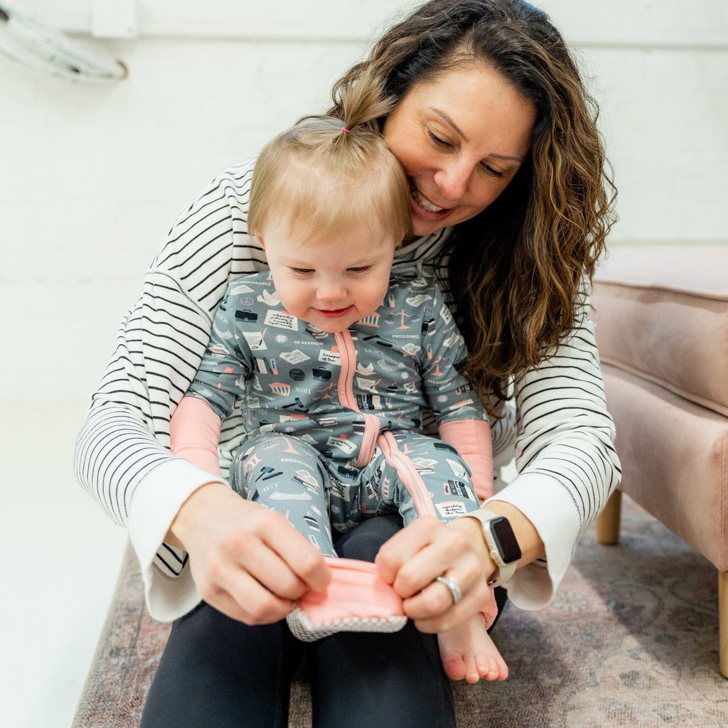 Order in the Cot Jams a smiling mother with long curly brown hair sits on the floor, holding a young girl on her lap. The girl, with light brown hair in a small ponytail, wears a grey footie pajama featuring law-themed illustrations, such as scales of justice, gavels, law books, and courthouse buildings. The pajama has light pink cuffs and a light pink two-way zipper. The mother, in a white and black striped long-sleeve shirt and black pants, helps her put on the foot covering.