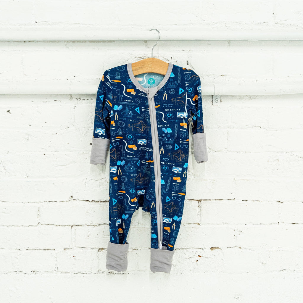 Roborest Jams navy blue baby footie pajama hangs on a wooden hanger against a white brick wall, featuring a detailed engineering, science, and technology-themed pattern. The deep navy background displays orange cranes, white light bulbs, gray gears, blue circuit boards, beakers with blue liquid, robotic arms, compasses, rulers, microscopes, wires, TV screens, atom symbols, and rocket ships. White text reads BUILD, CREATE, DEVELOP, and DREAM. The pajama has a two-way zipper, light gray trim, and cuffs.