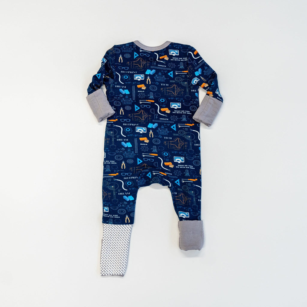 Roborest Jams back view of a navy blue baby footie pajama showcases a vibrant engineering, science, and technology-themed pattern. The pajama features images like orange cranes, white light bulbs, gray gears, circuit boards, beakers with blue liquid, robotic arms, compasses, rulers, microscopes, wires, TV screens, atom symbols, and rocket ships. White text reads BUILD,CREATE, DEVELOP, and DREAM. The pajama has light gray cuffs and fold-over footies, with anti-slip grippy dots on the left foot.