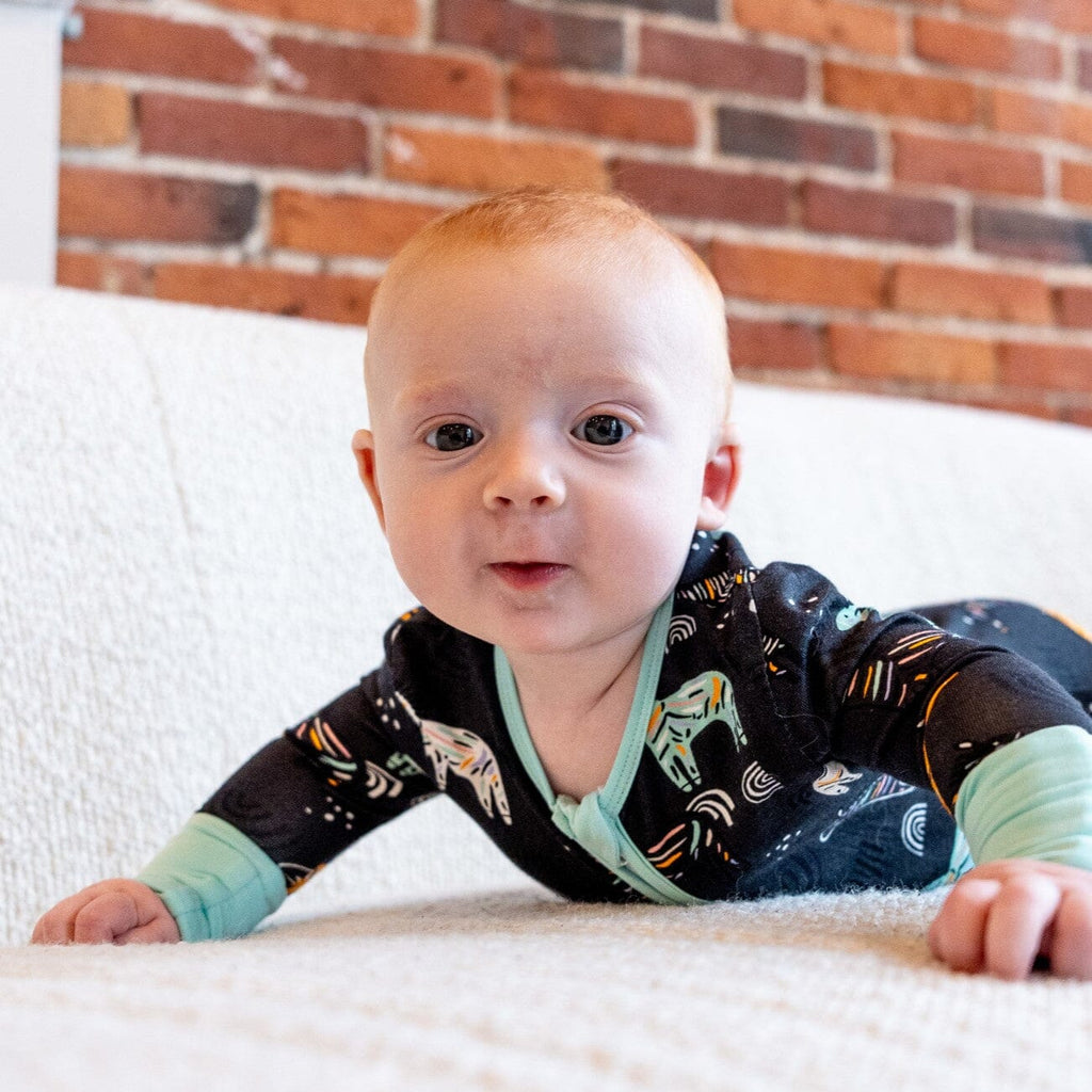 Zzzebra a baby boy with light hair is lying on his stomach on a white textured couch. He is dressed in black footie pajamas with a two-way zipper and light blue cuffs. The pajamas feature a colorful pattern of zebras and rainbows in light blue, orange, and white. The baby boy is looking up with a curious expression and there is a red brick wall in the background.