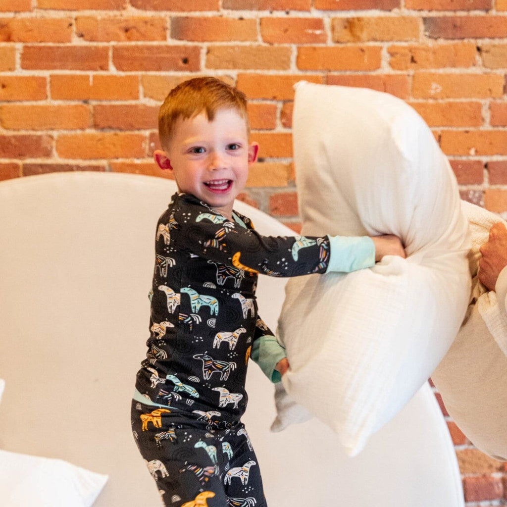 Zzzebra Big Kids Jams a boy with short ginger hair is in a pillow fight, holding a large white pillow. He is wearing a black long-sleeved bamboo pajamas with light blue cuffs and colorful zebras and rainbows. The boy is looking towards the camera with a wide smile. 