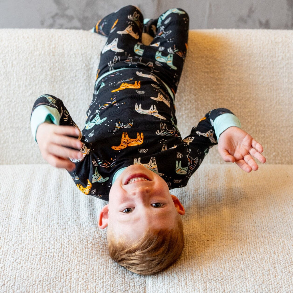 Zzzebra Big Kids Jams a young boy with light brown hair is lying upside down on a white textured couch. He is wearing a black long-sleeved bamboo pajama set with light blue cuffs. The pajamas feature a colorful pattern of zebras and rainbows in light blue, orange, and white. The boy is smiling widely, with his head hanging over the edge of the couch.