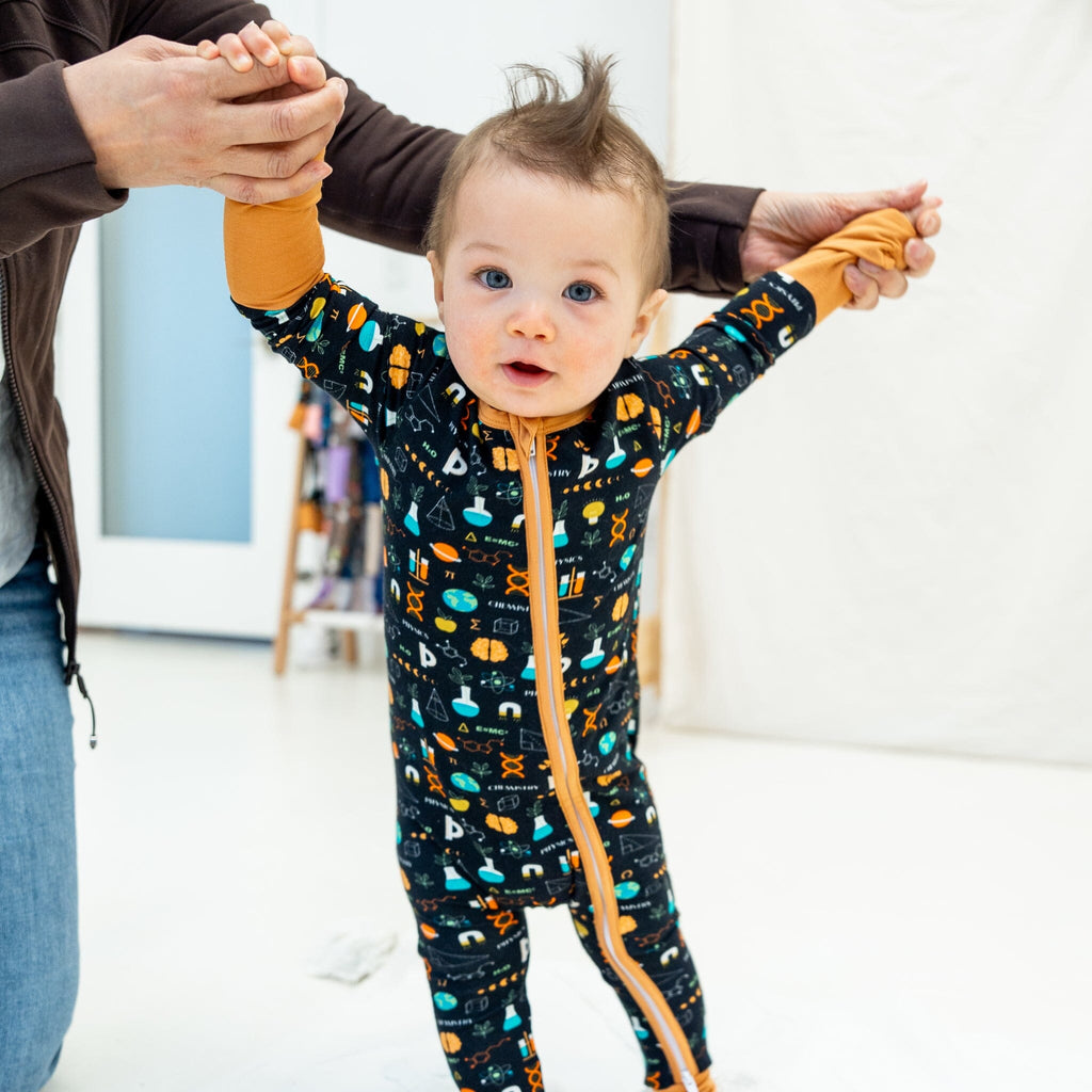 Keep An Ion Bedtime Jams a toddler boy with light brown hair styled in a small mohawk stands with a curious expression, holding his mother's hands for support. He wears a black baby footie pajama with a vibrant science-themed pattern, featuring DNA helixes, atoms, microscopes, flasks, test tubes, equations, books, 'Physics' and 'Chemistry' text, and icons like planet earth, a ring planet, and light bulbs in orange, blue, green, and white. The pajama has a two-way zipper and mustard yellow cuffs. 