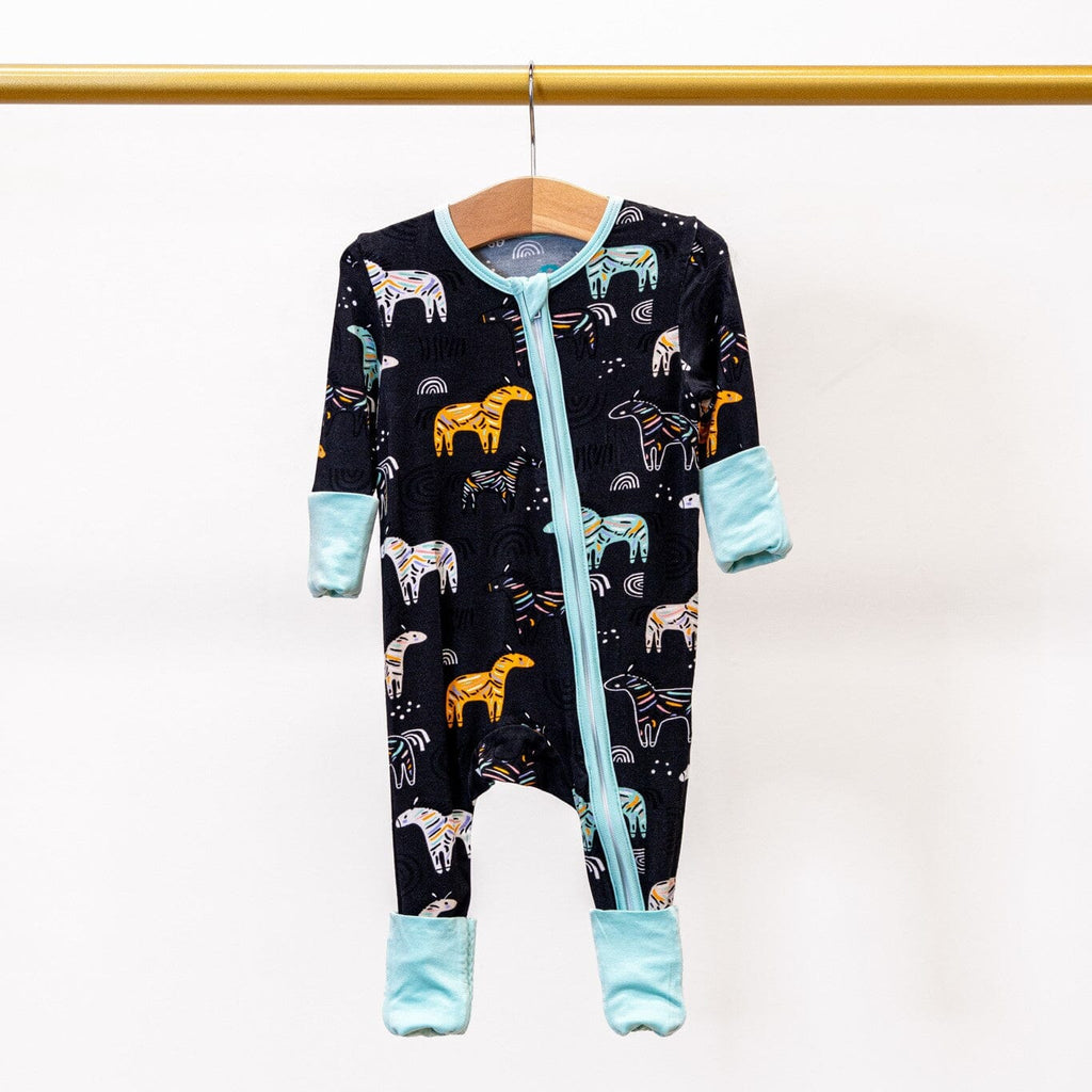 Zzzebra Black footie bamboo pajamas with a two way zipper with a colorful zebra and rainbow print featuring turquoise trim and cuffs, hanging on a wooden hanger.