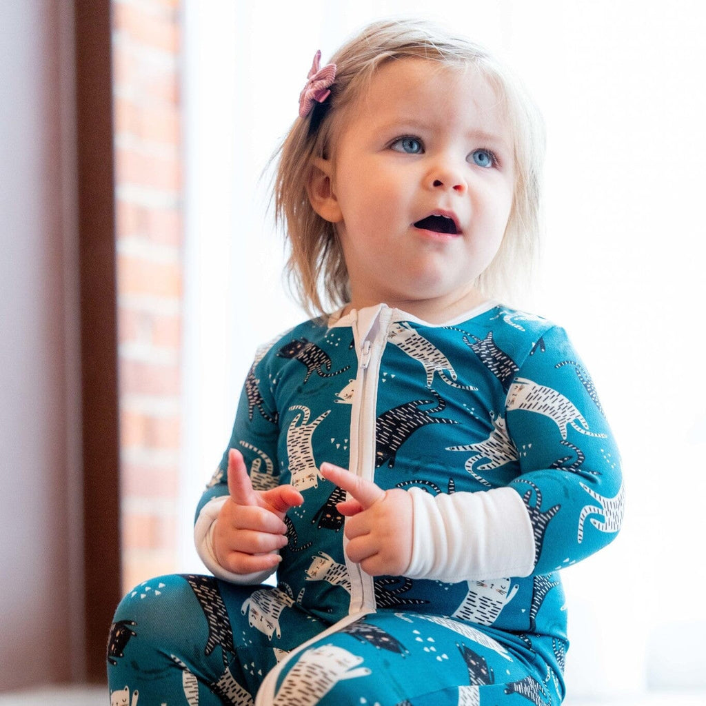 Paws and Relax toddler sitting wearing teal footie bamboo pajamas with a two-way zipper and a pattern of cats in white and black. She has a pink hair clip and is looking up with a curious expression