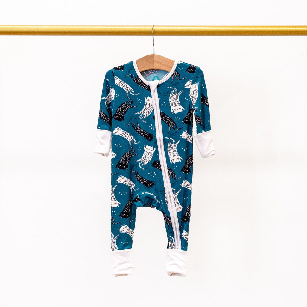 Paws and Relax teal-colored footie pajamas with a two-way zipper, featuring a pattern of white and black cats featuring white trim and cuffs, hanging on a wooden hanger against a plain white background.