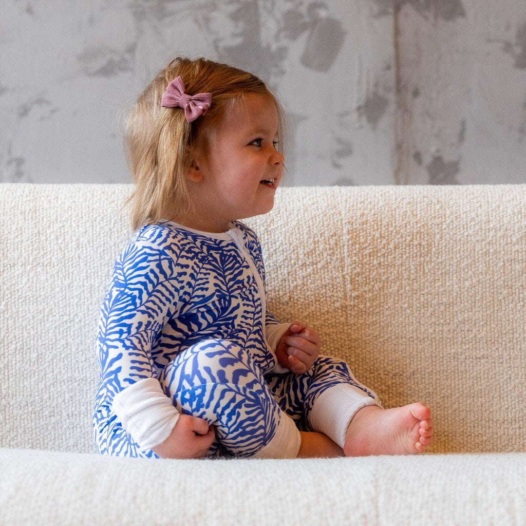 Seabreeze a toddler with light brown hair, partially held back with a pink bow, is sitting sideways on a white textured couch. She is wearing a footie pajamas with a two-way zipper with a blue and white fern-like pattern.The girl is smiling and looking to her left, showing her profile. Her feet are bare, and she is sitting with her knees bent, holding her legs close to her body.