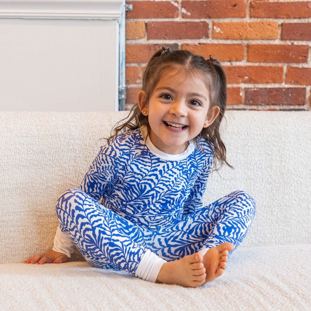 Seabreeze Big Kids Jams a smiling girl with brown hair in pigtails sitting cross-legged on a white textured couch. She is wearing a long-sleeved bamboo pajama top and matching bamboo pajama pants with a blue and white fern-like pattern.