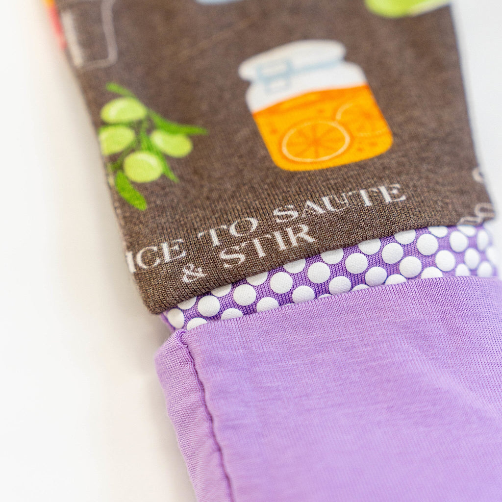 Dreams a la Mode Jams a close-up view of the grippy foot of a baby’s footie pajamas. The fabric features a brown background with colorful illustrations of kitchen items, including a jar with an orange slice and green olives. The sole has a lavender background with white grippy dots, and the edge of the foot is lavender.