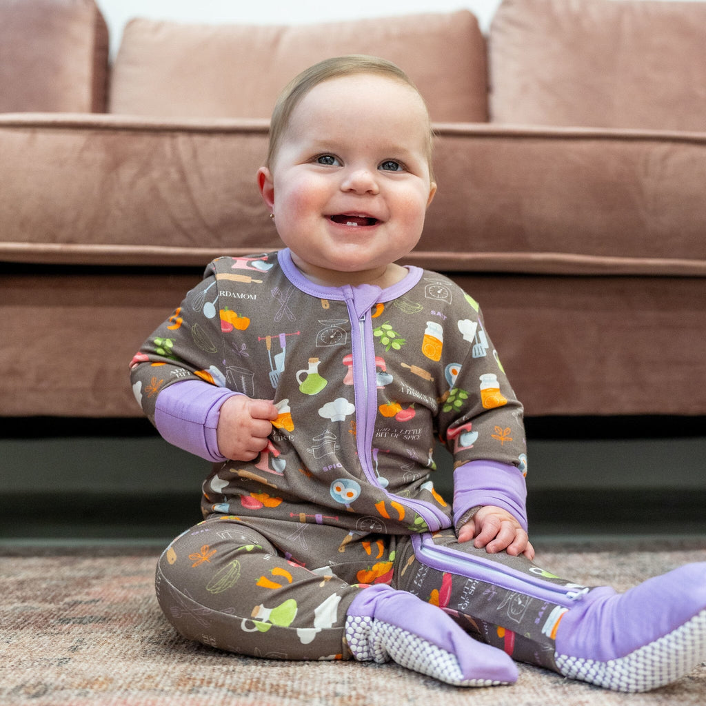 Dreams a la Mode Jams a smiling baby boy with light brown hair sits on a patterned rug with a pink couch in the background. He wears a brown footie pajama with a cooking-themed pattern featuring spatulas, whisks, measuring spoons, forks, pots, a red mixer, a rolling pin, bell peppers, eggplants, carrots, olives, two eggs in a pan, a chef's hat, and salt shakers. The pajama has a two-way zipper, lavender trim and cuffs, white non-slip grippies, and text like 'salt' and 'cinnamon.