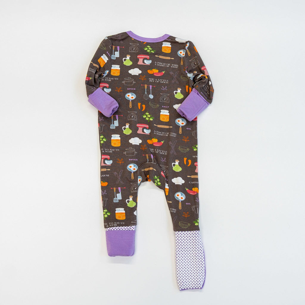 Dreams a la Mode Jams the back view of a brown baby footie pajama with a colorful kitchen-themed pattern featuring spatulas, knives, whisks, measuring spoons, forks, pots, a red mixer, a rolling pin, bell peppers, eggplants, carrots, olives, two eggs in a pan, a chef's hat, and salt shakers. The pajama has lavender cuffs, with the left foot showing a lavender fold-over footie, and the right foot showing anti-slip grippy dots.