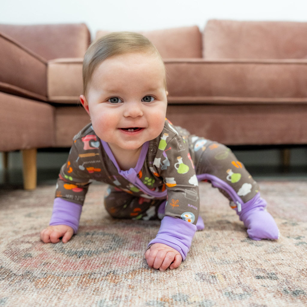 Dreams a la Mode Jams a baby boy smiling with light brown hair is crawling on a patterned rug, with a pink couch in the background. The baby is wearing footie pajamas with a two-way zipper in a brown fabric featuring colorful kitchen-themed illustrations, such as vegetables, pots, and utensils in shades of green, orange, and white. The pajamas have lavender cuffs and fold-over fotties. 