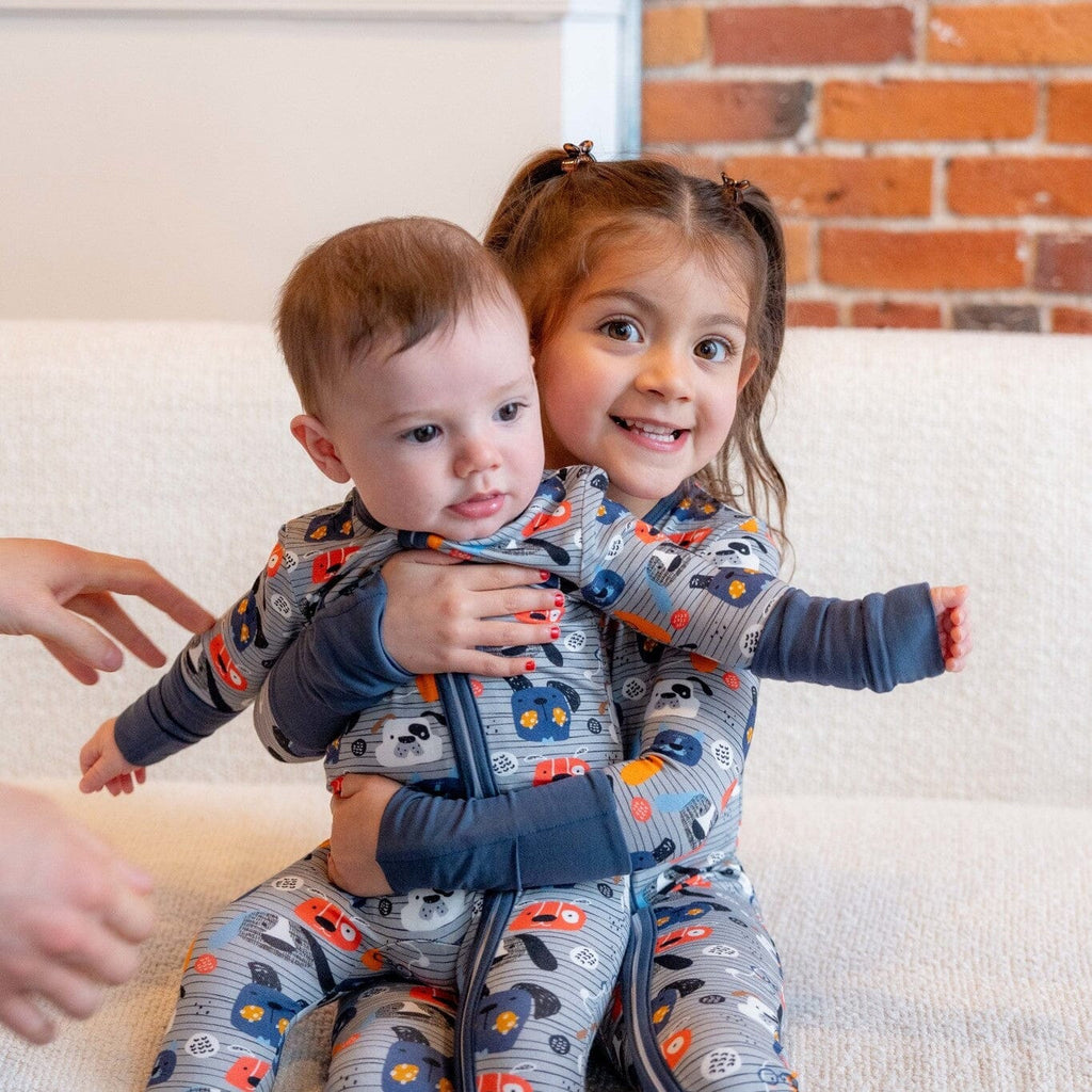 Paws and Snooze a girl with brown curly hair in pigtails sitting on a white couch, holding a baby in her lap. Both children are wearing matching grey footie bamboo pajamas with a two-way zipper and a  pattern of colorful dog faces in orange, blue, and white. An adult's hands are seen reaching towards the baby. The background has a red brick wall.