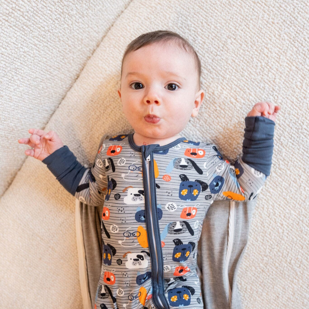 Paws and Snooze a baby lying on a beige textured surface and an opened sleepsack, wearing grey footie bamboo pajamas with a two-way zipper and a pattern of colorful dog faces in orange, blue, and white. The baby has a playful expression  with puckered lips