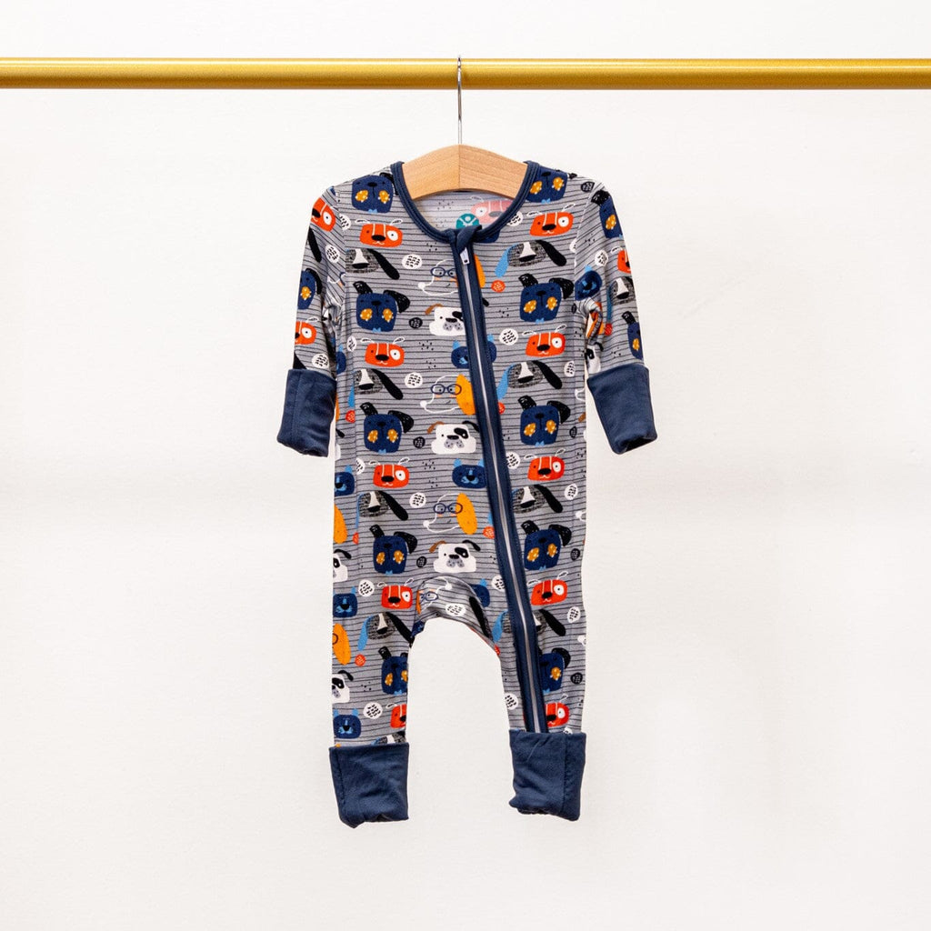 Paws and Snooze a grey footie pajama with a pattern of colorful dog faces in orange, blue, and white. The pajama has a two-way zipper with dark blue trim and cuffs, and it is hanging on a wooden hanger from a gold-colored rod against a plain white background.