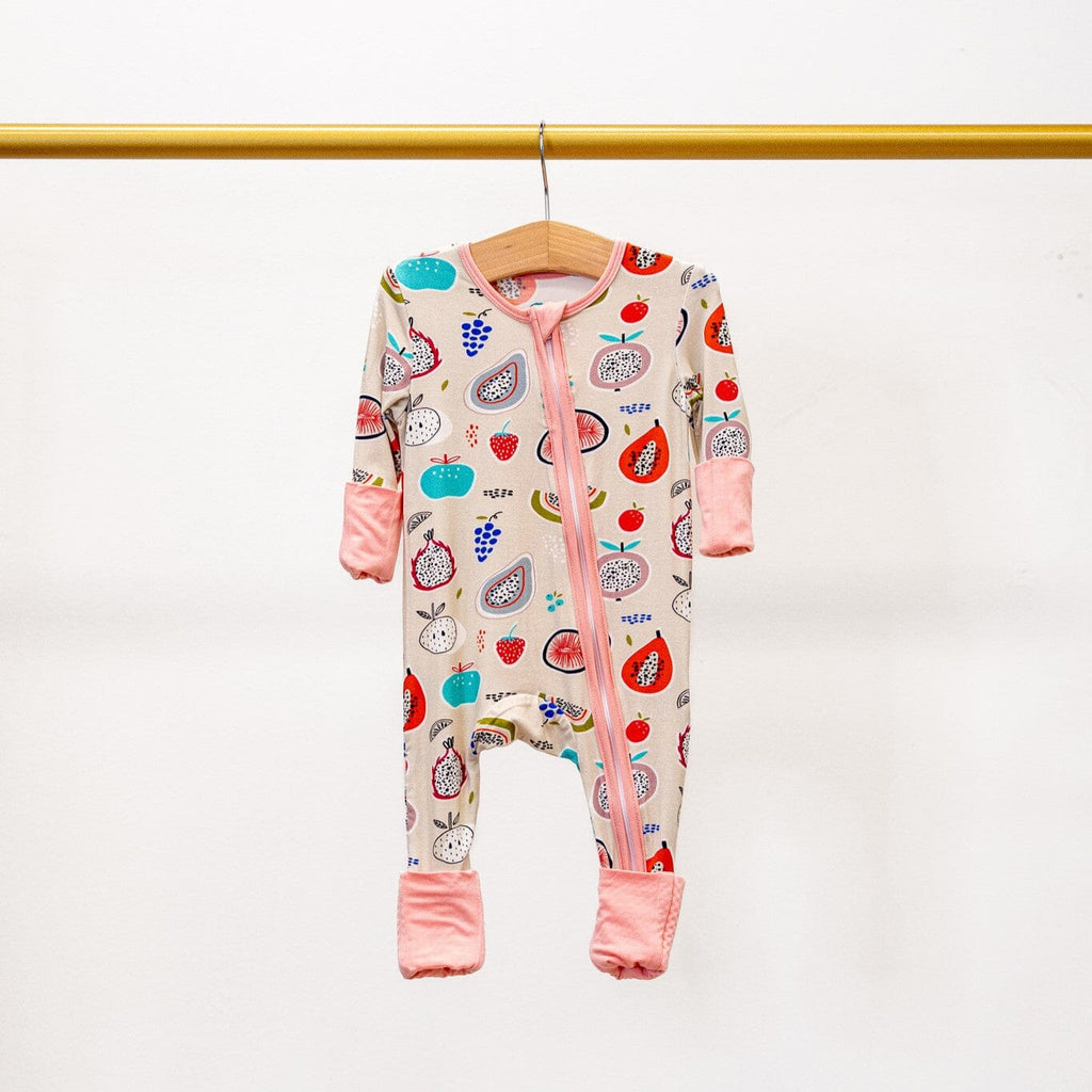 Kiwi Kisses light pink/cream footie pajama with a colorful fruit pattern featuring apples, strawberries, pomegranates, dragon fruits, grapes, and blueberries. The pajama has a two-way zipper, pink trim, and cuffs, and is hanging on a wooden hanger from a gold-colored rod against a plain white background.