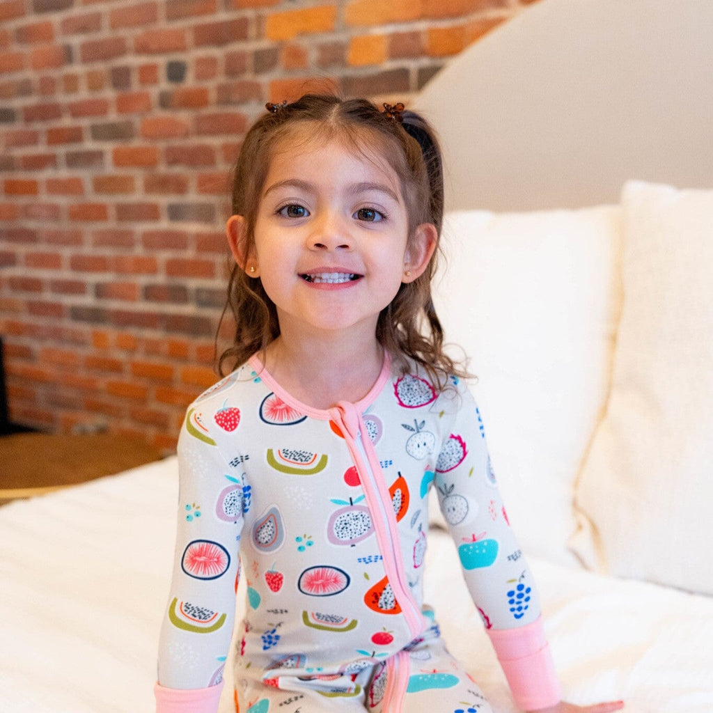 Kiwi Kisses a smiling girl with brown hair in pigtails wearing bamboo footie pajamas with a two-way zipper decorated with colorful fruit patterns sits on a bed with  white bedding and a brick wall background.