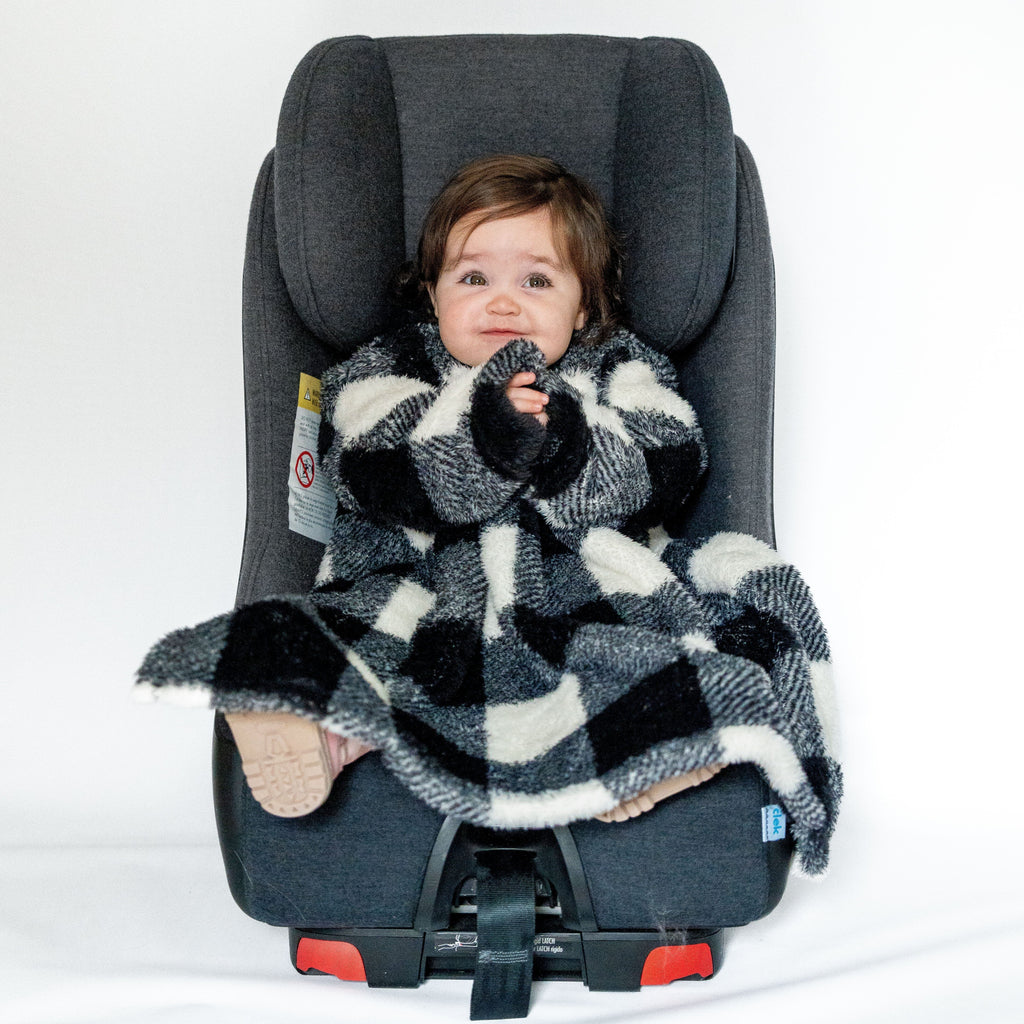 A toddler with light brown hair is sitting in a grey car seat. The child is wearing a Car Seat Blankie in a black and white plaid pattern. The blanket has long sleeves and covers the child up to their feet, which are wearing beige rubber-soled shoes. The toddler has their hands near their mouth and is looking ahead with a calm expression. The background is plain white.