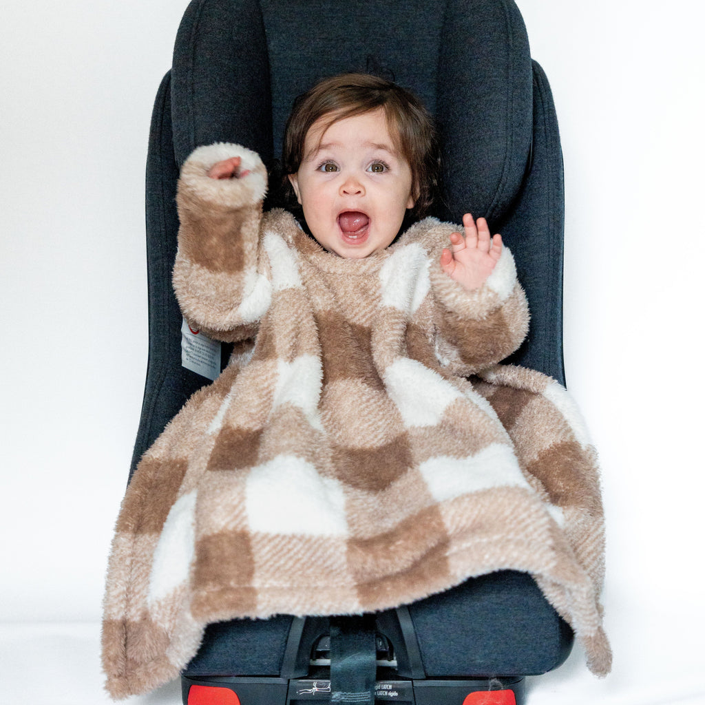 A baby sits in a car seat, wearing a cozy car seat blankie with a soft, brown and white checkered pattern. The baby has a happy expression, with arms raised, and the car seat is black, set against a plain white background