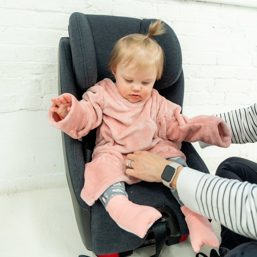 A toddler with blonde hair in a small ponytail, dressed in a pink velvet car seat blankie, sits in a dark gray car seat while the mother's hand that is wearing a striped shirt and a smartwatch is adjusting the blankie. The blankie has a plush texture, with wide sleeves and attached foot covers. The child also wears gray footie pajama under the blankie. The background is a white brick wall.