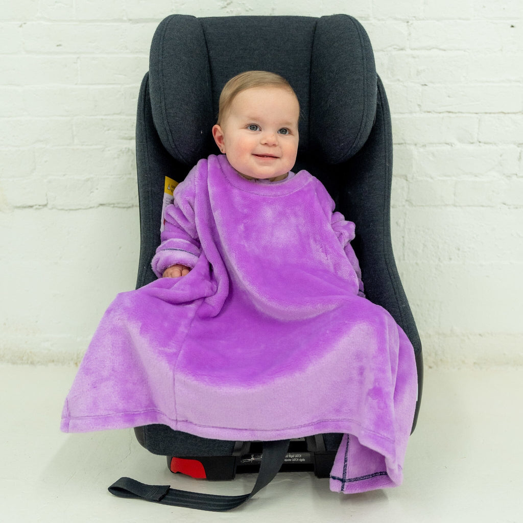 A toddler with light brown hair, dressed in a purple velvet car seat blankie, sits in a dark gray car seat. The blankie has a plush texture and wide sleeves, enveloping the child in its soft fabric. The child smiles happily. The background is a white brick wall