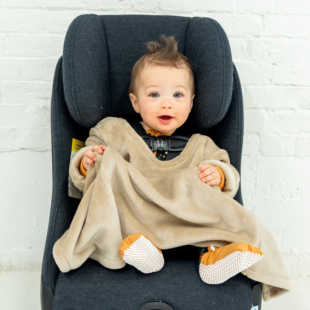 A smiling baby boy sitting in a car seat, wearing a beige velvet-textured Car Seat Blankie. He has light brown hair styled in a small mohawk, blue eyes, with hands outstretched. The Car Seat Blankie covers the baby's legs and has orange cuffs and his feet is covered with anti-slip grippy footie. The car seat is dark grey and set against a white brick wall.