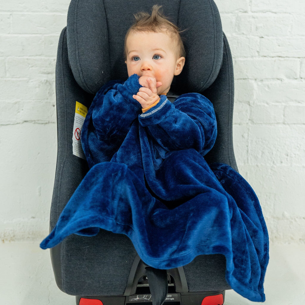 A boy toddler with light brown hair styled into a small tuft at the top sits in a dark gray car seat, dressed in a deep blue velvet car seat blankie. The blankie has a soft, plush texture, with wide sleeves and a cozy design. He is sucking his hands while gazing off to the side. The background is a white brick wall.