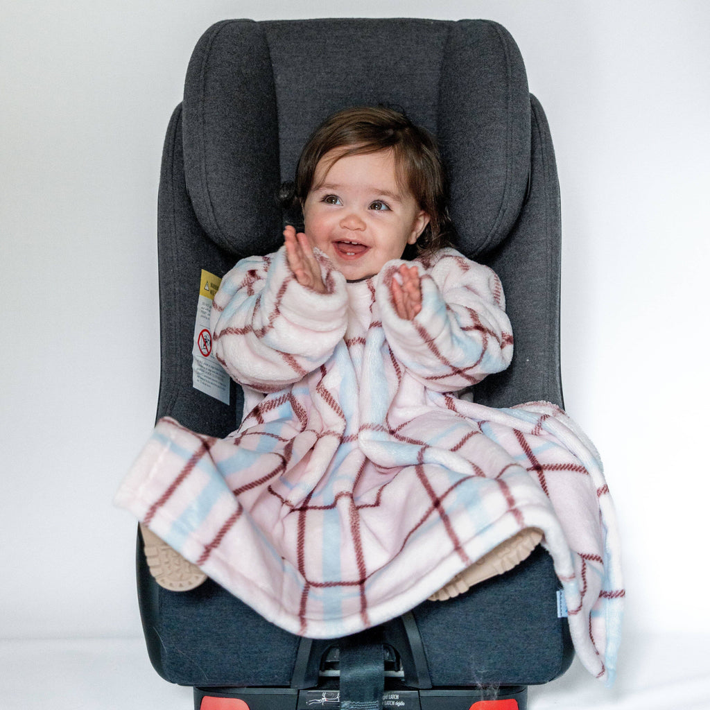 A toddler is seated in a dark gray car seat, wearing a cozy pink and blue plaid car seat blankie. The baby is smiling and clapping hands, with the car seat positioned against a plain white background.