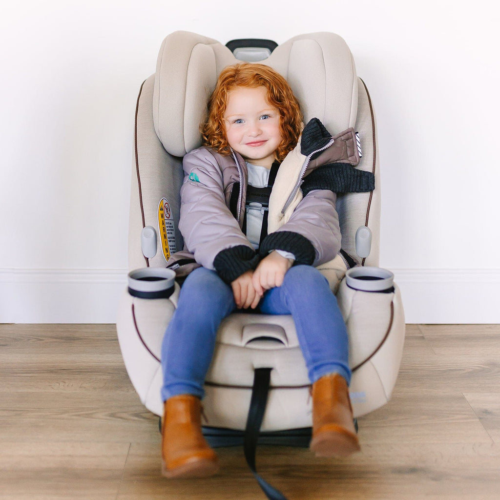 The Adventurer/Brown/Grey
Little girl with red curly hair and blue eyes is wearing a grey car seat coat. She is sitting in a beige car seat with the coat unzipped showing the buckled harness.